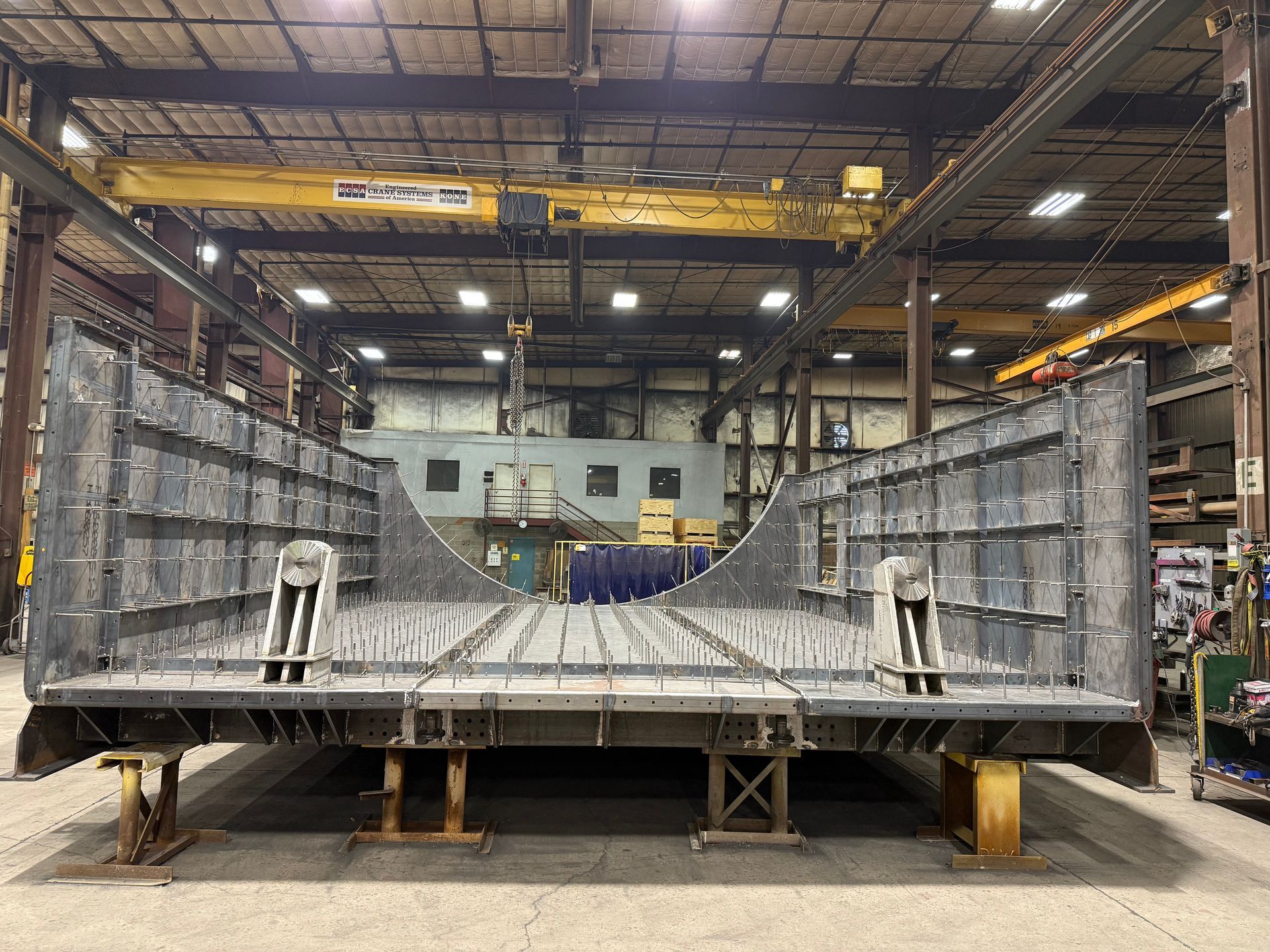 Large, gray metal structure in a workshop, possibly a ship hull section, supported by wooden blocks.