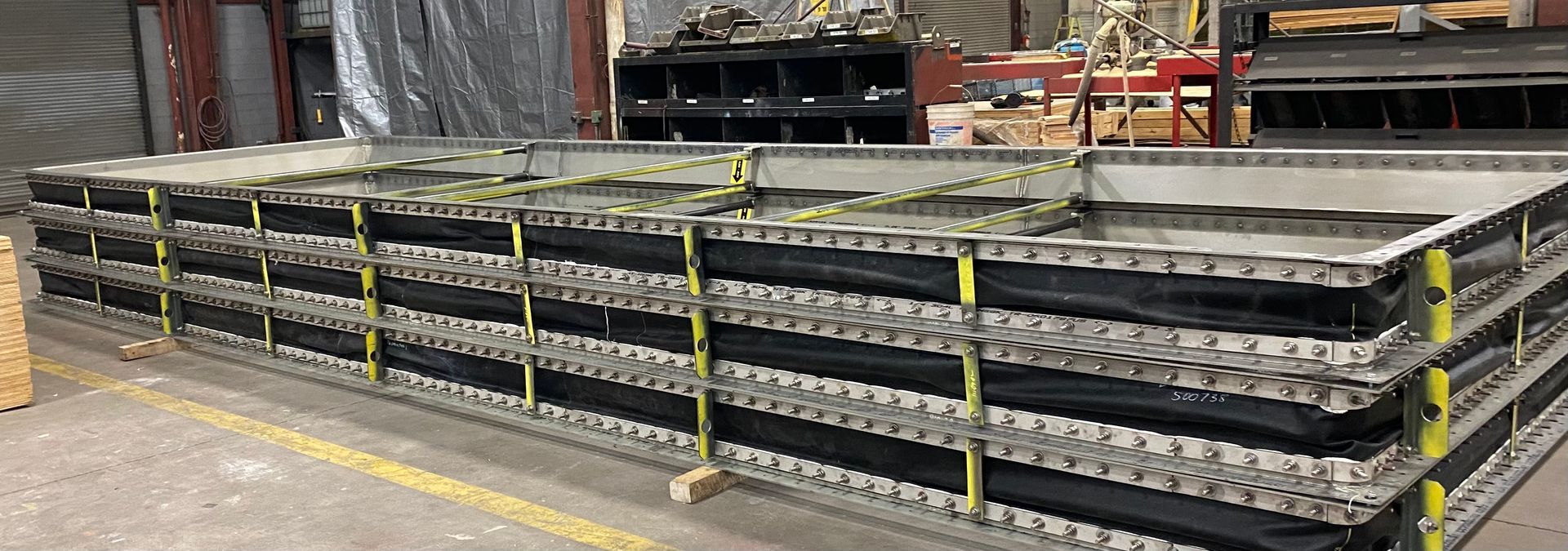Stacked rectangular metal panels, secured with black straps and yellow bands, in an industrial setting.