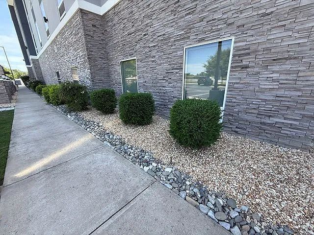 A sidewalk leading to a building with a stone wall and bushes in front of it.