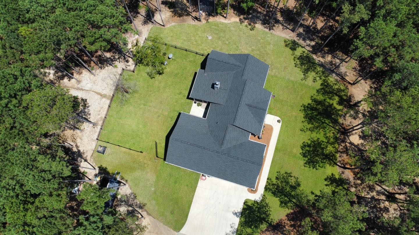 Aerial view of a house with a dark roof, surrounded by green grass and pine trees.