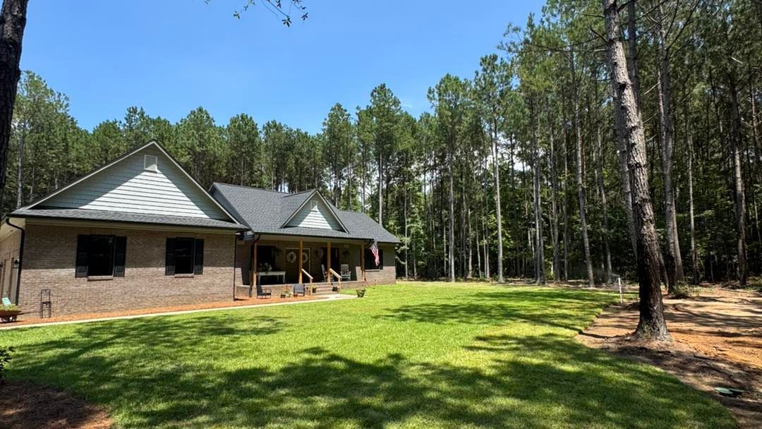 A one-story brick house with a porch and green lawn, surrounded by trees on a sunny day.