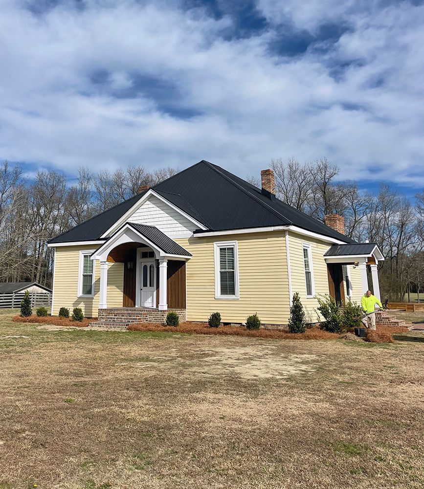 A man is standing in front of a yellow house with a black roof.