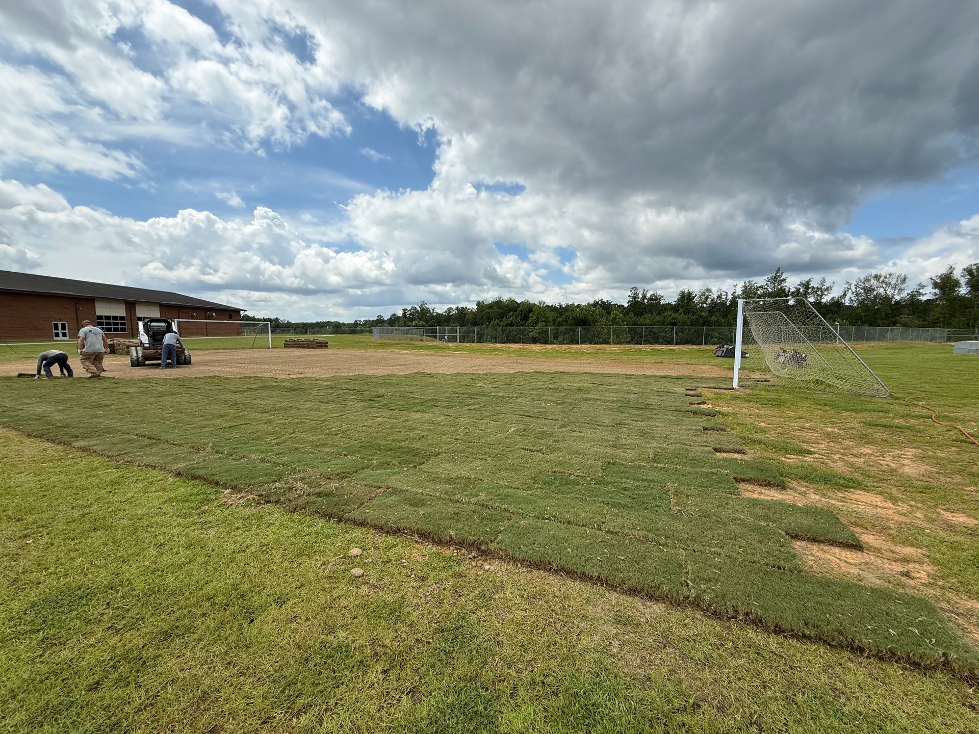 Workers laying sod on a grassy field, with a school building and soccer goal visible under a cloudy sky.