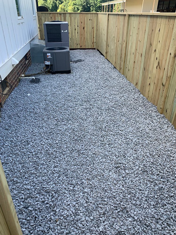 A gravel patio with a wooden fence and an air conditioner.