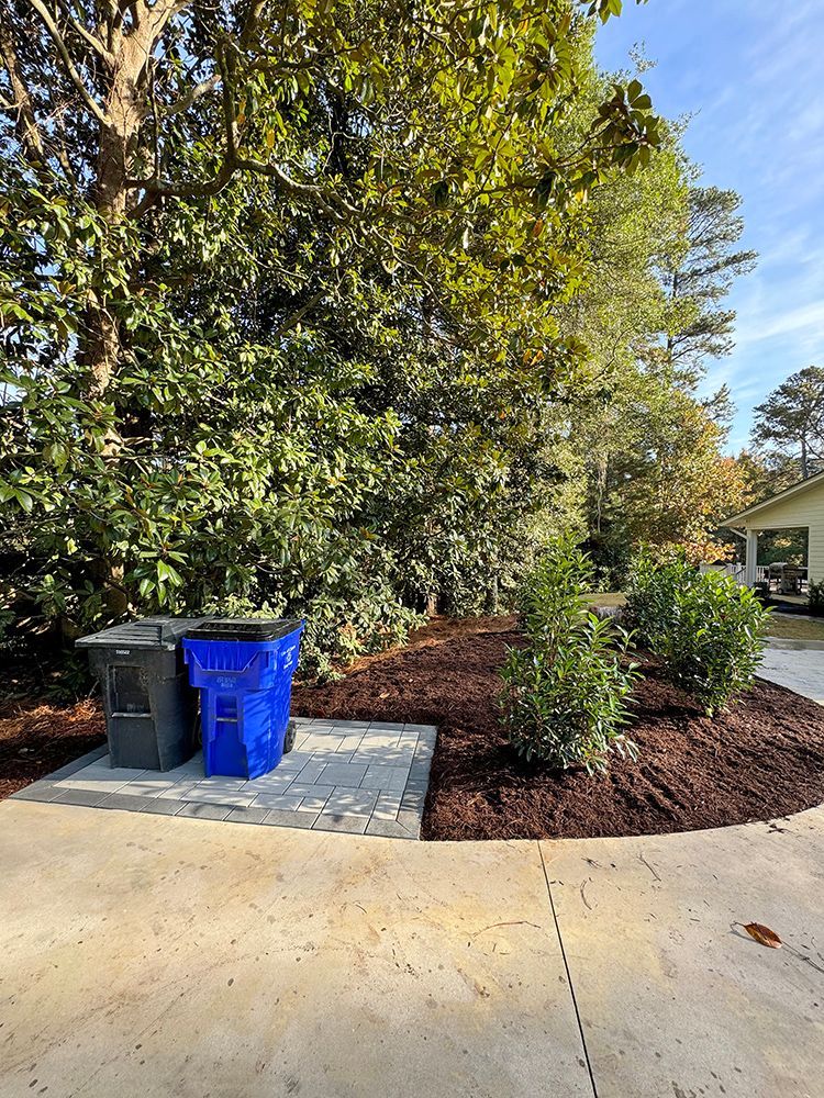 A blue trash can is sitting in the middle of a driveway next to a tree.