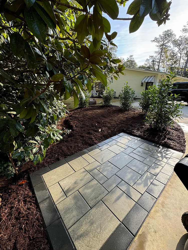 A brick walkway leading to a house surrounded by trees and mulch.