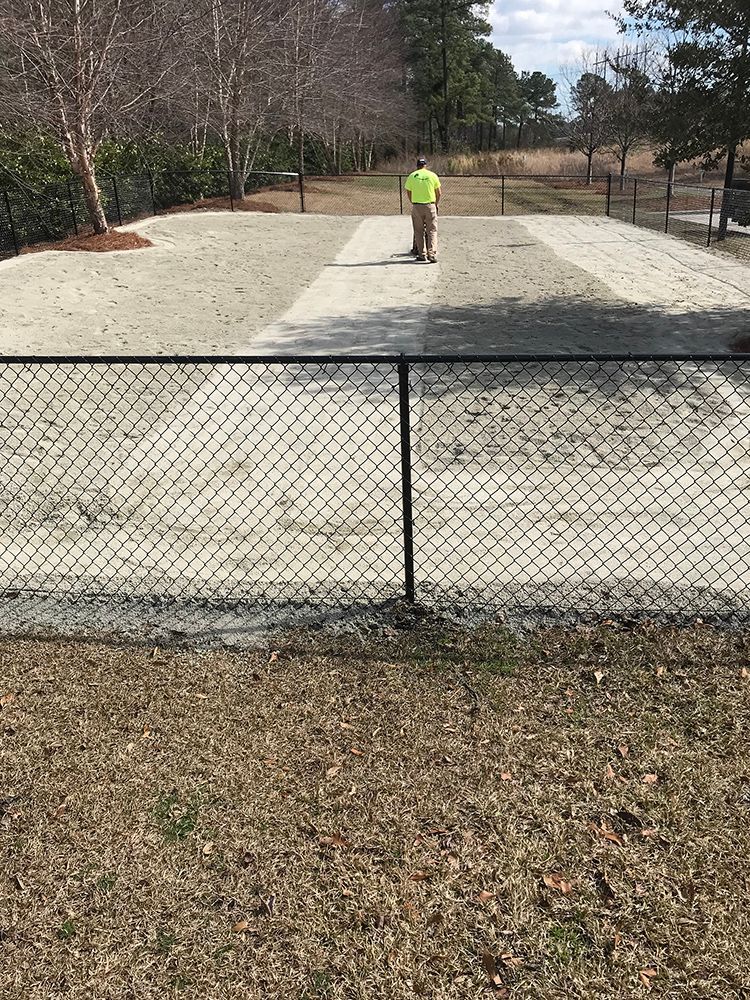 A man is walking down a dirt road next to a chain link fence.