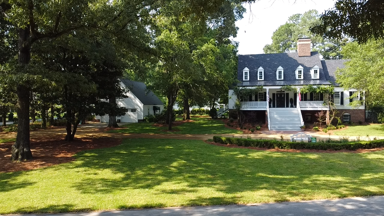 A large white house with a black roof is surrounded by trees and grass.