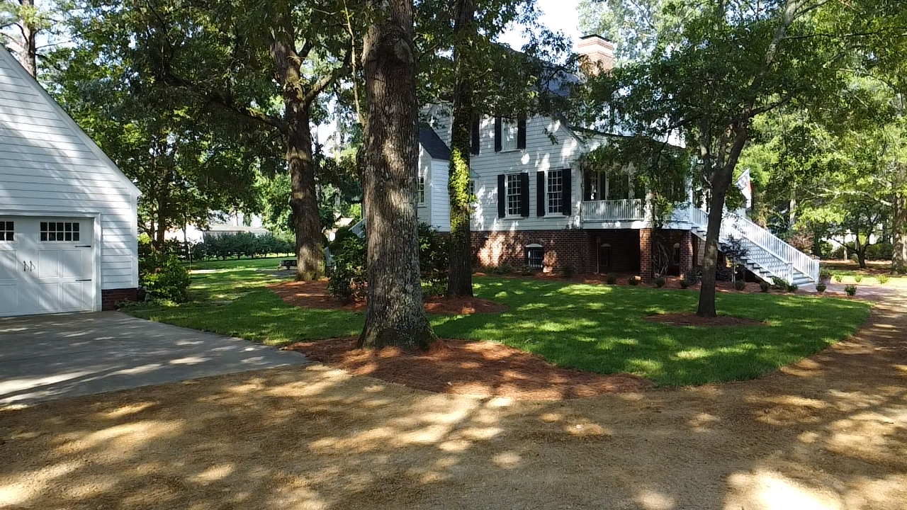 A large white house is surrounded by trees and a gravel driveway.
