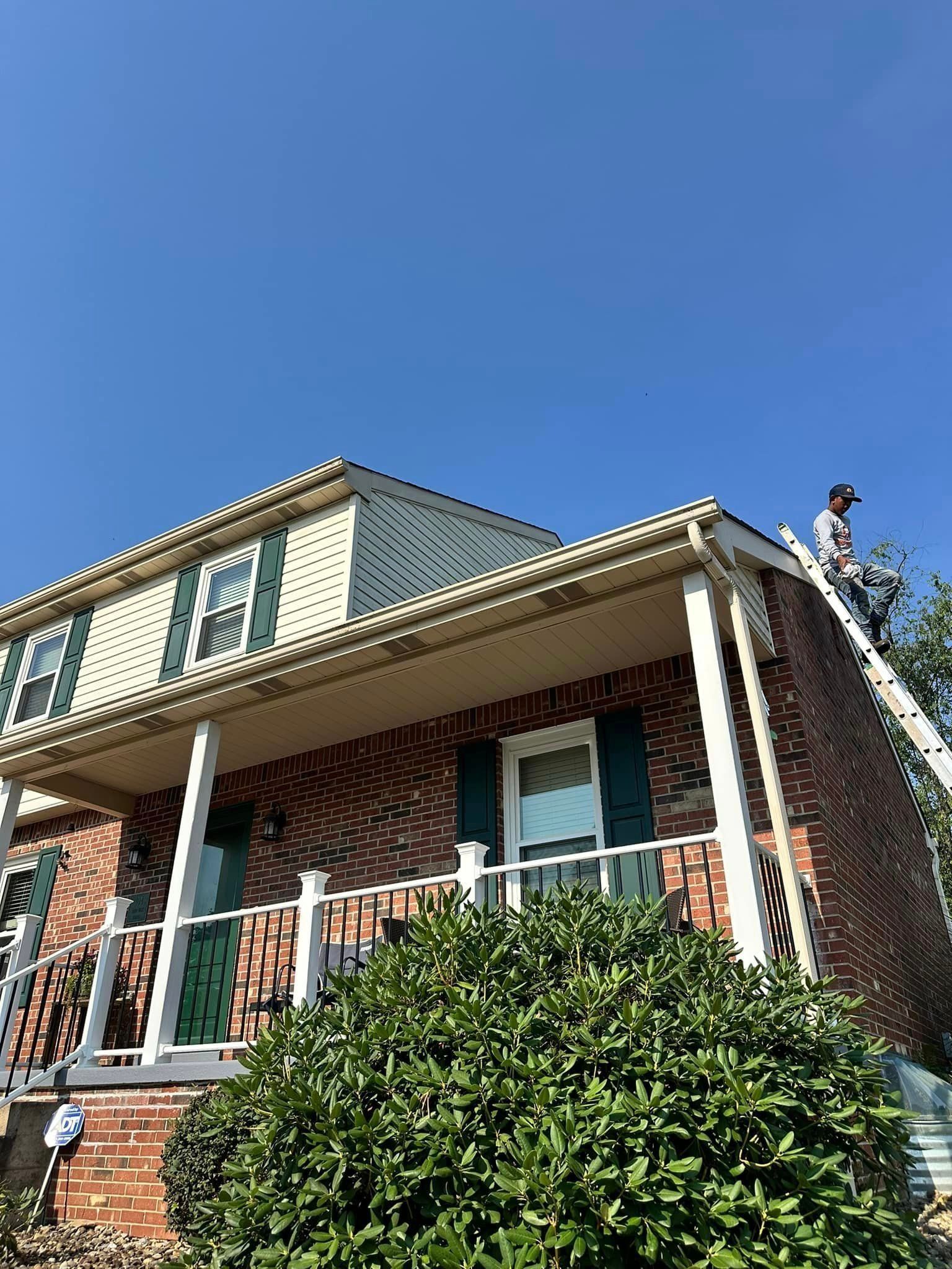 A person on a ladder working on the gutters of a two-story brick house with green shutters under a blue sky.
