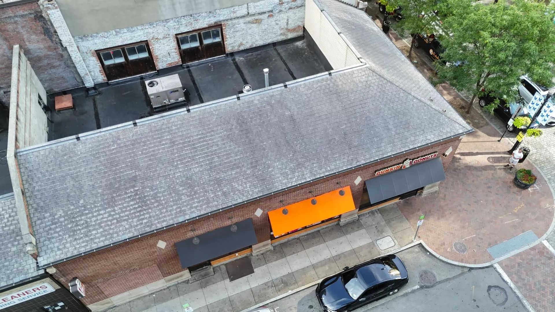 Aerial view of a brick building with awnings, a black car parked nearby, and a small tree on a sidewalk.