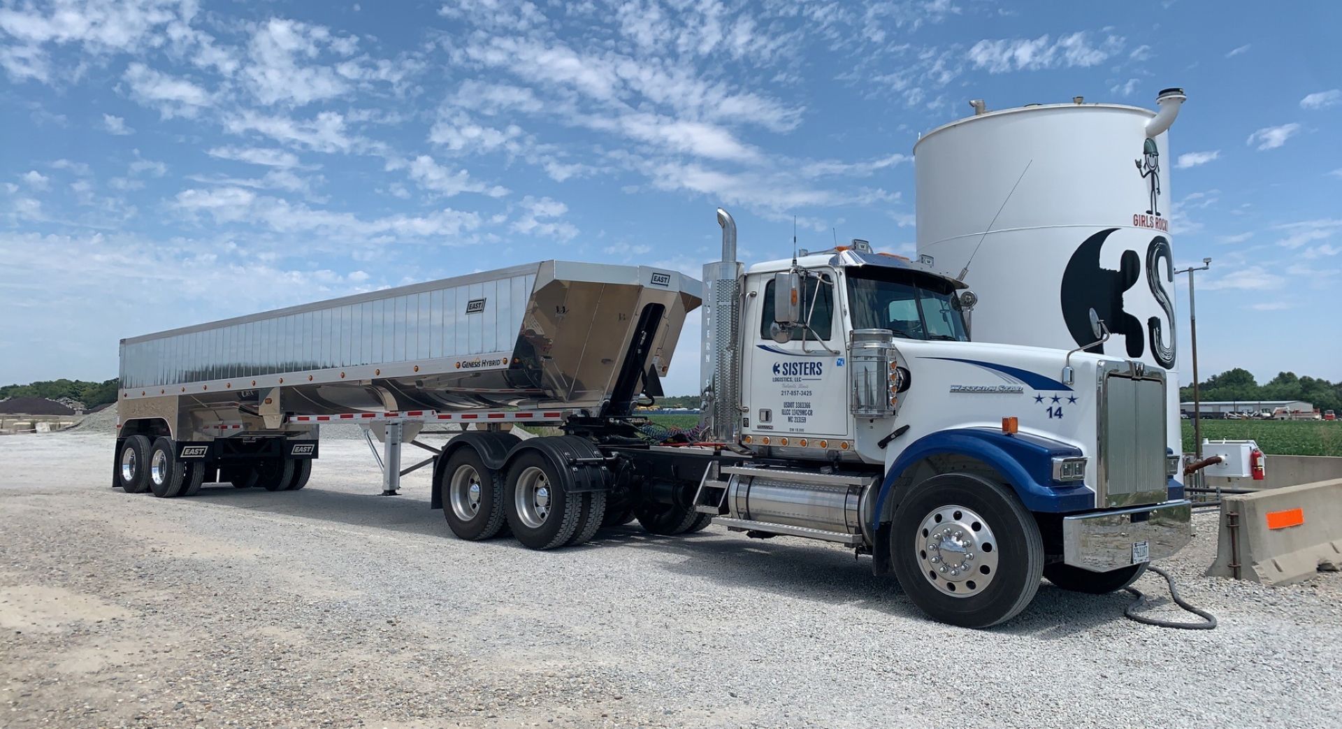 White semi truck with silver trailer parked on gravel under a blue sky.