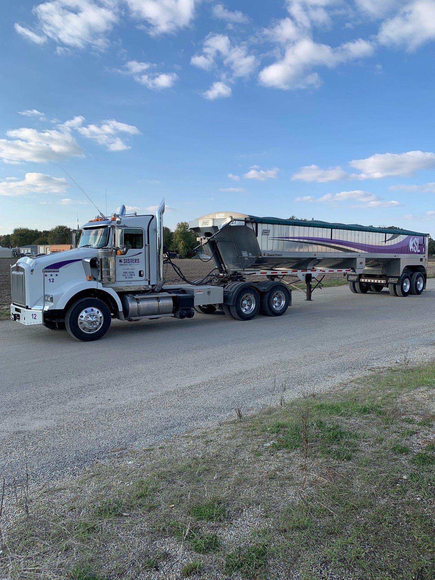 White semi-truck hauling a loaded dump trailer on a gravel road under a blue, partly cloudy sky