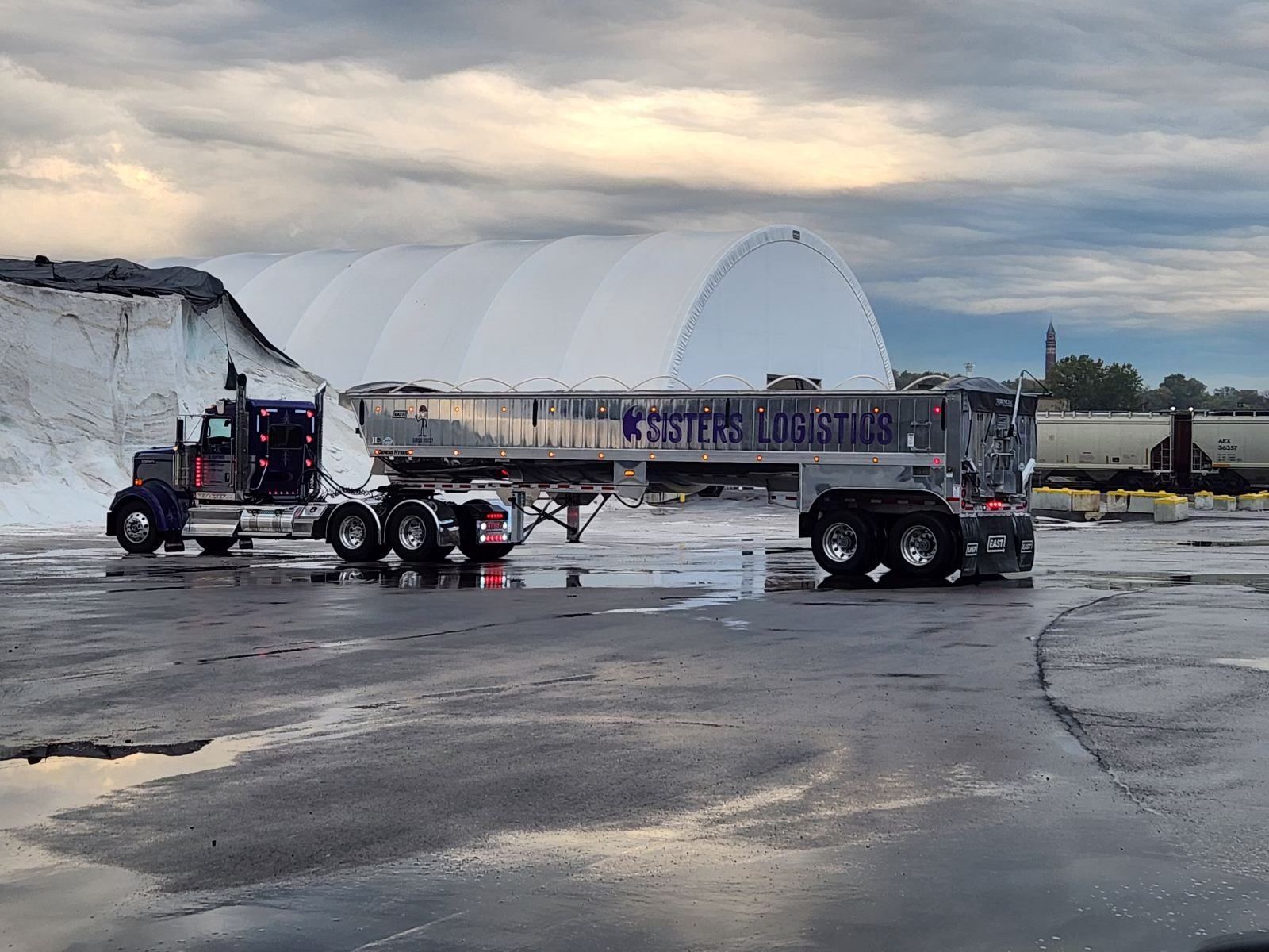 Truck hauling a long trailer in an industrial lot with snow piles and puddles under cloudy skies