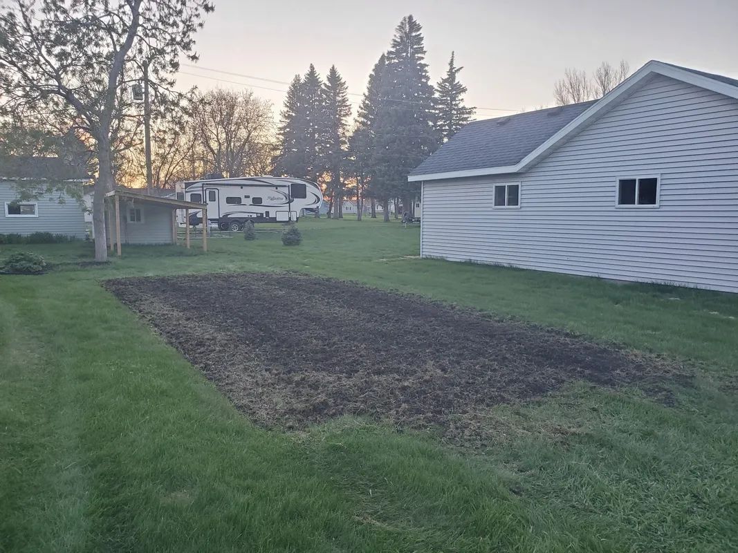 A backyard with a tilled garden bed, a white house, and an RV parked in the distance.