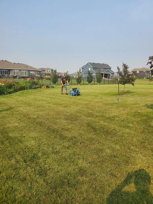 Man mowing a large green lawn in a suburban backyard under a blue sky.