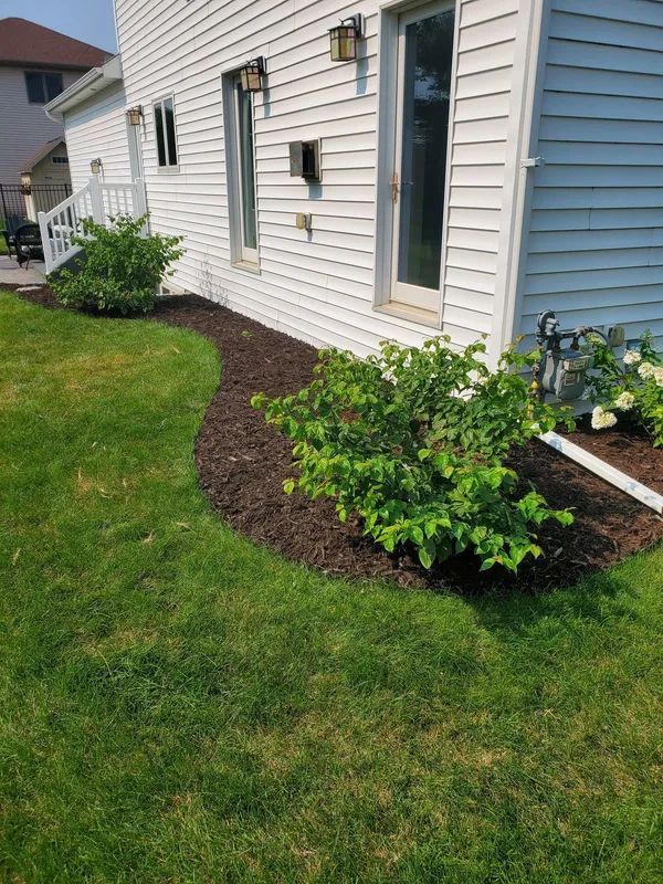 Green lawn borders a house with tan siding and a mulch bed with green bushes.