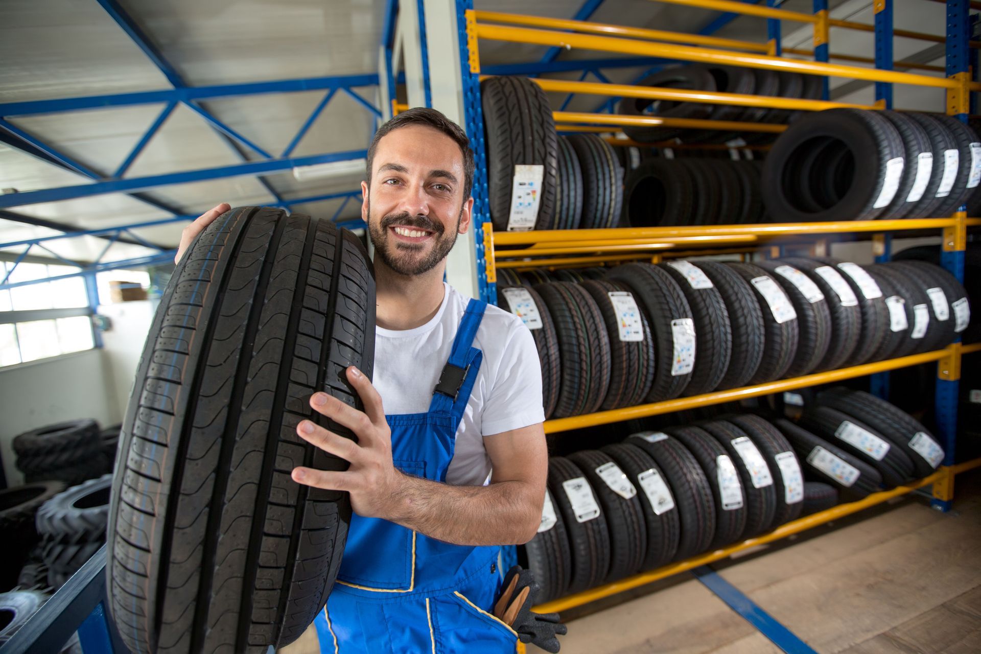 Mechanic holding a tire, smiling, in a tire shop. Yellow shelves filled with tires in background.