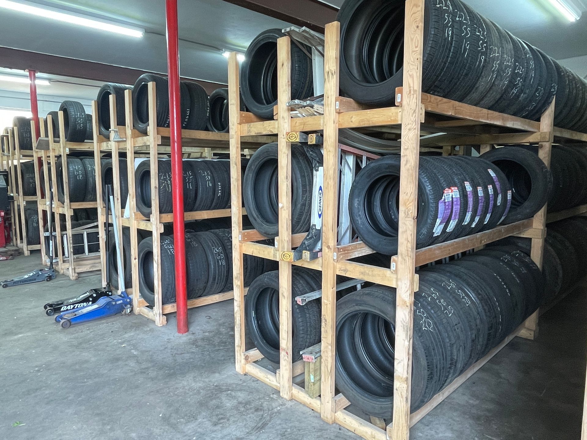 Rows of black tires neatly stacked on wooden warehouse storage shelves under bright ceiling lights.