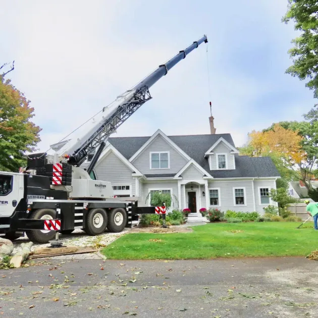 Crane truck lifting equipment near a gray house with a front porch and autumn trees
