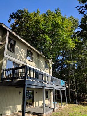 Two-story beige house with dark balcony, set among tall green trees on a sunny day