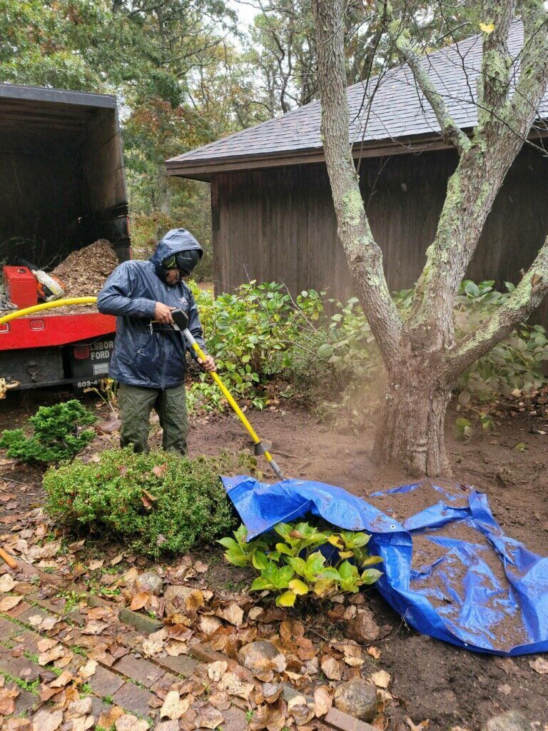 Person in rain jacket using a yellow trimmer to clear weeds by a tree and blue tarp in a wooded yard.