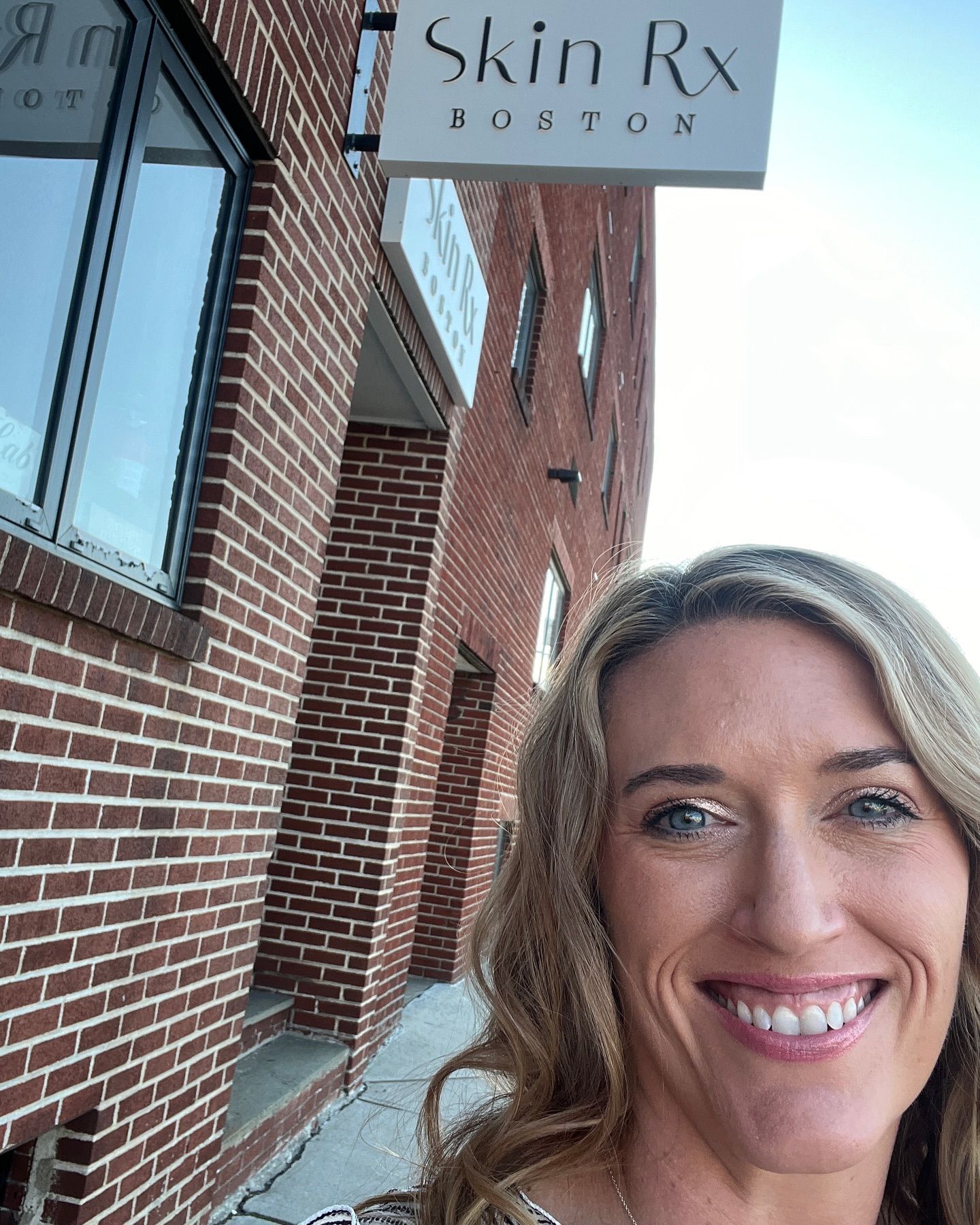 A woman is smiling in front of a brick building with a sign that says skin rx