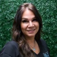 A woman in a black shirt is smiling in front of a green hedge.