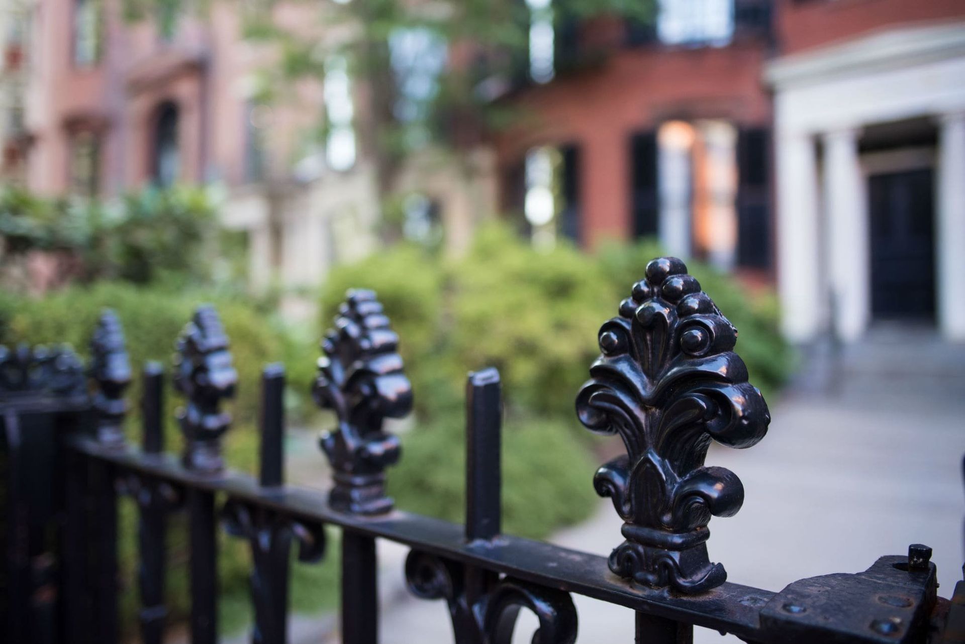 Black wrought-iron fence in front of brick buildings and greenery on a city street