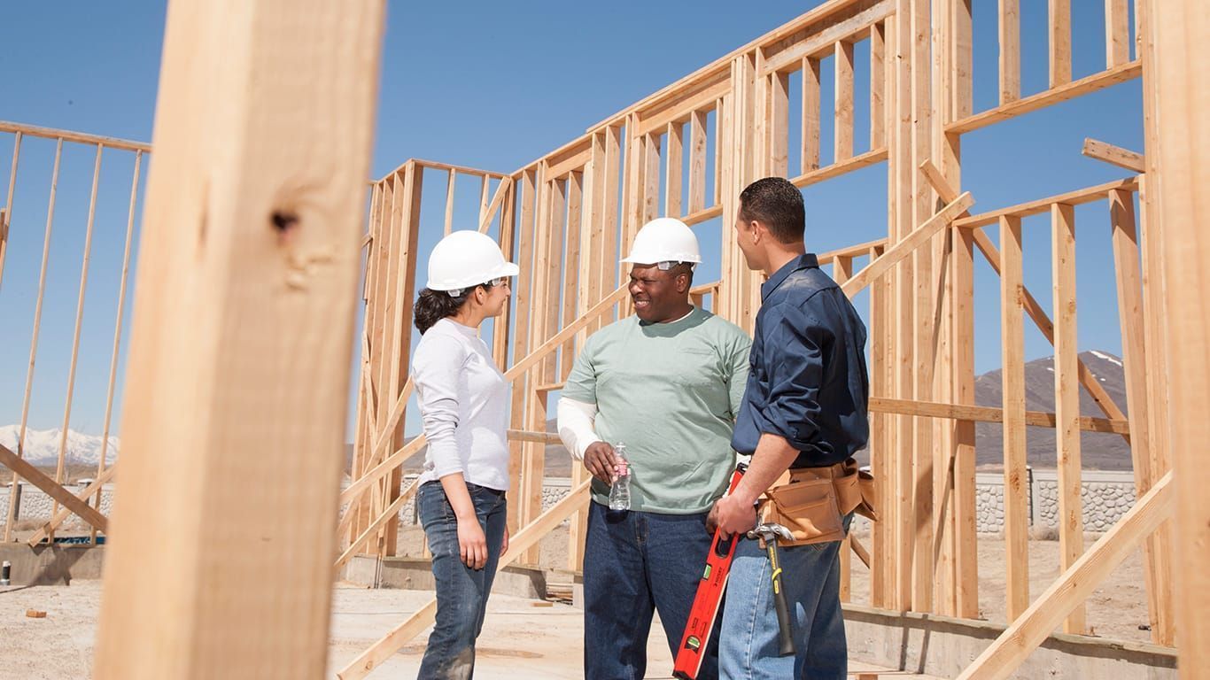 Three construction workers in hard hats discuss framing at a wooden house site.