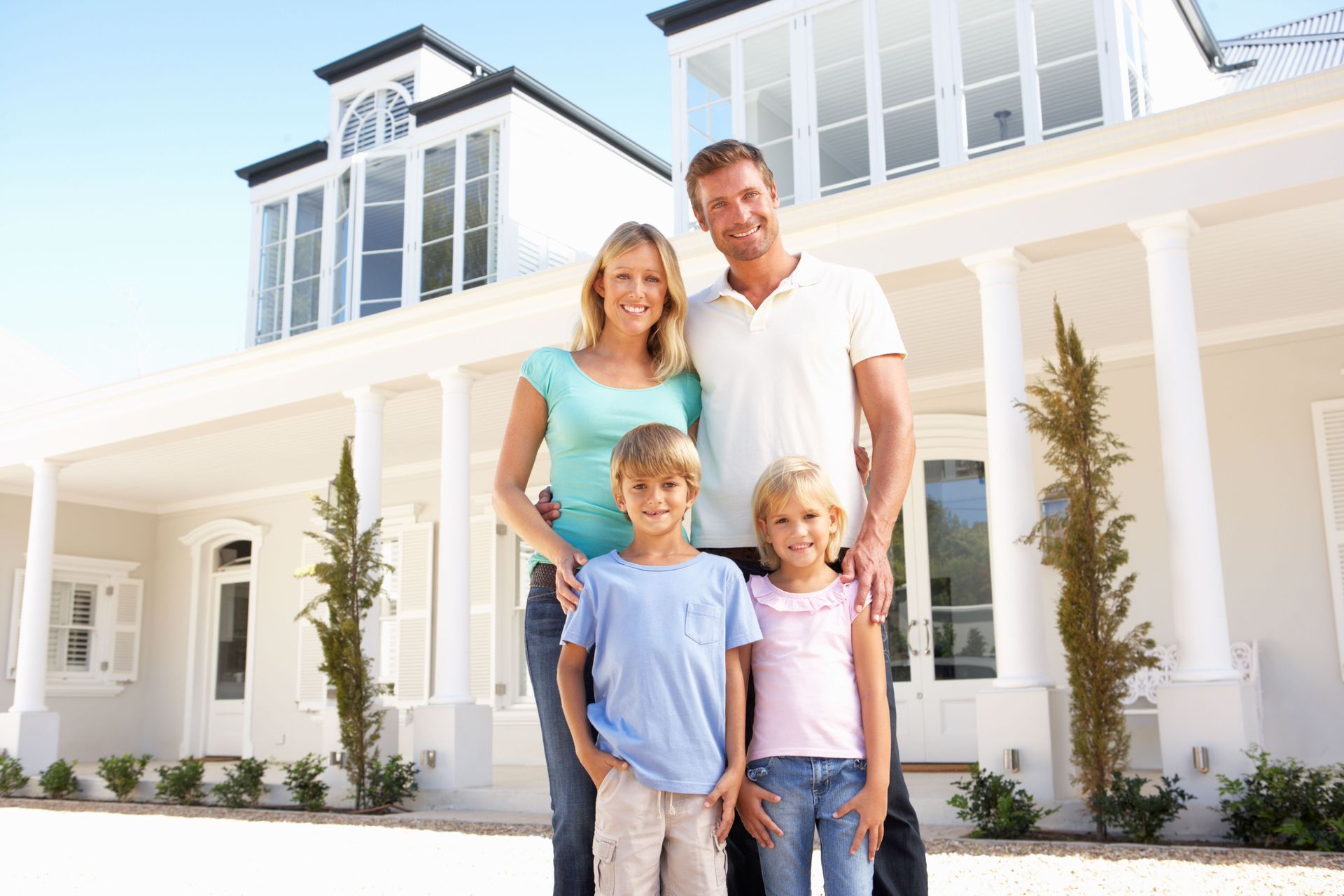 A family of four stands smiling in front of a white, multi-story house with columns.
