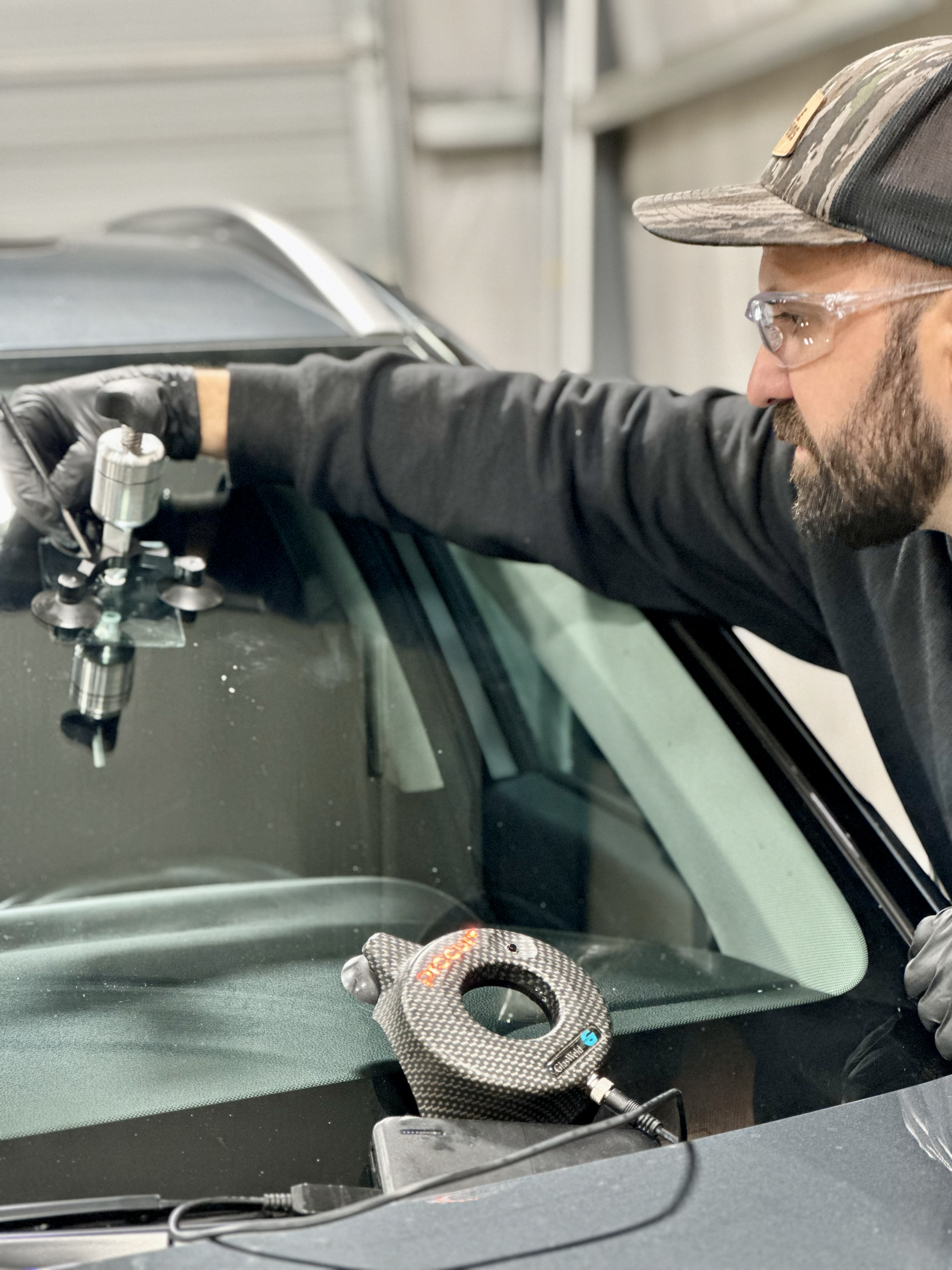 A person repairing a car windshield. They are holding tools, wearing glasses, and gloves, indoors.