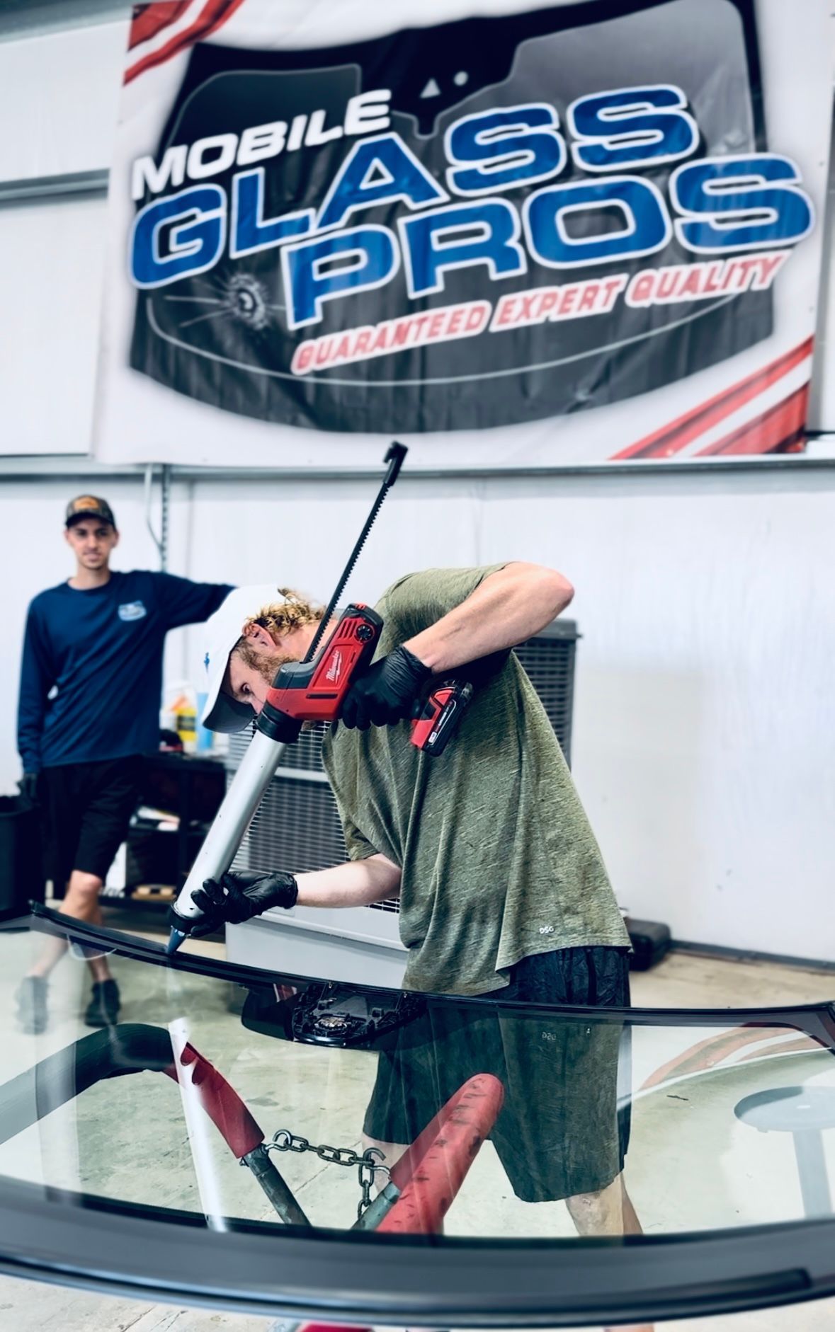 Man using a caulking gun on a car windshield, another man observes. "Mobile Glass Pros" sign in background.