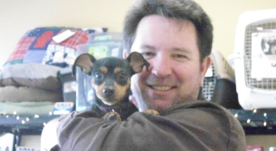 Man holding a small black and tan dog, both smiling. Interior setting with pet supplies.