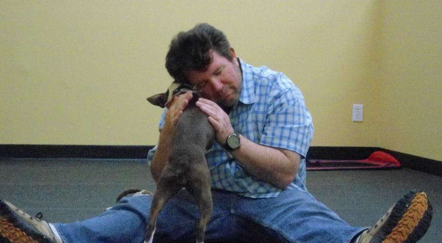 Man hugging a small dog, both sitting on floor, yellow wall in background.