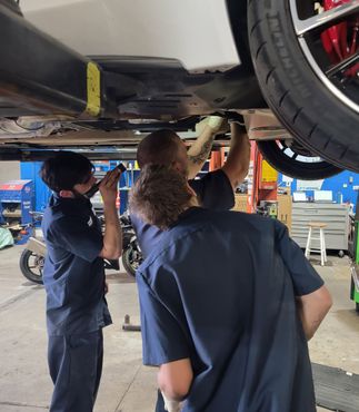 Three mechanics working under a car in a shop. One holds a flashlight, others are inspecting the underside.