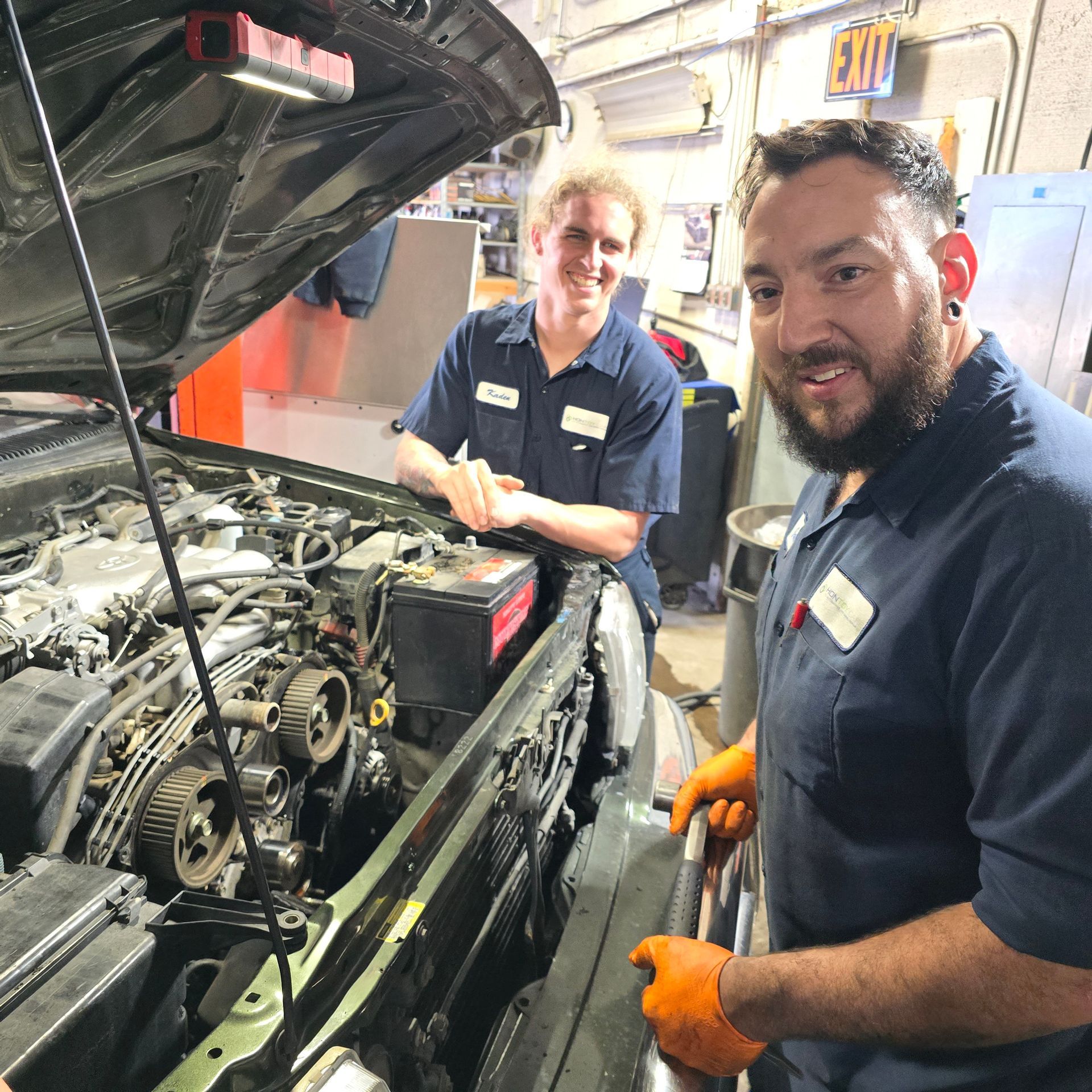 Two mechanics working on a car engine in a garage. One holds a tool, both smile.