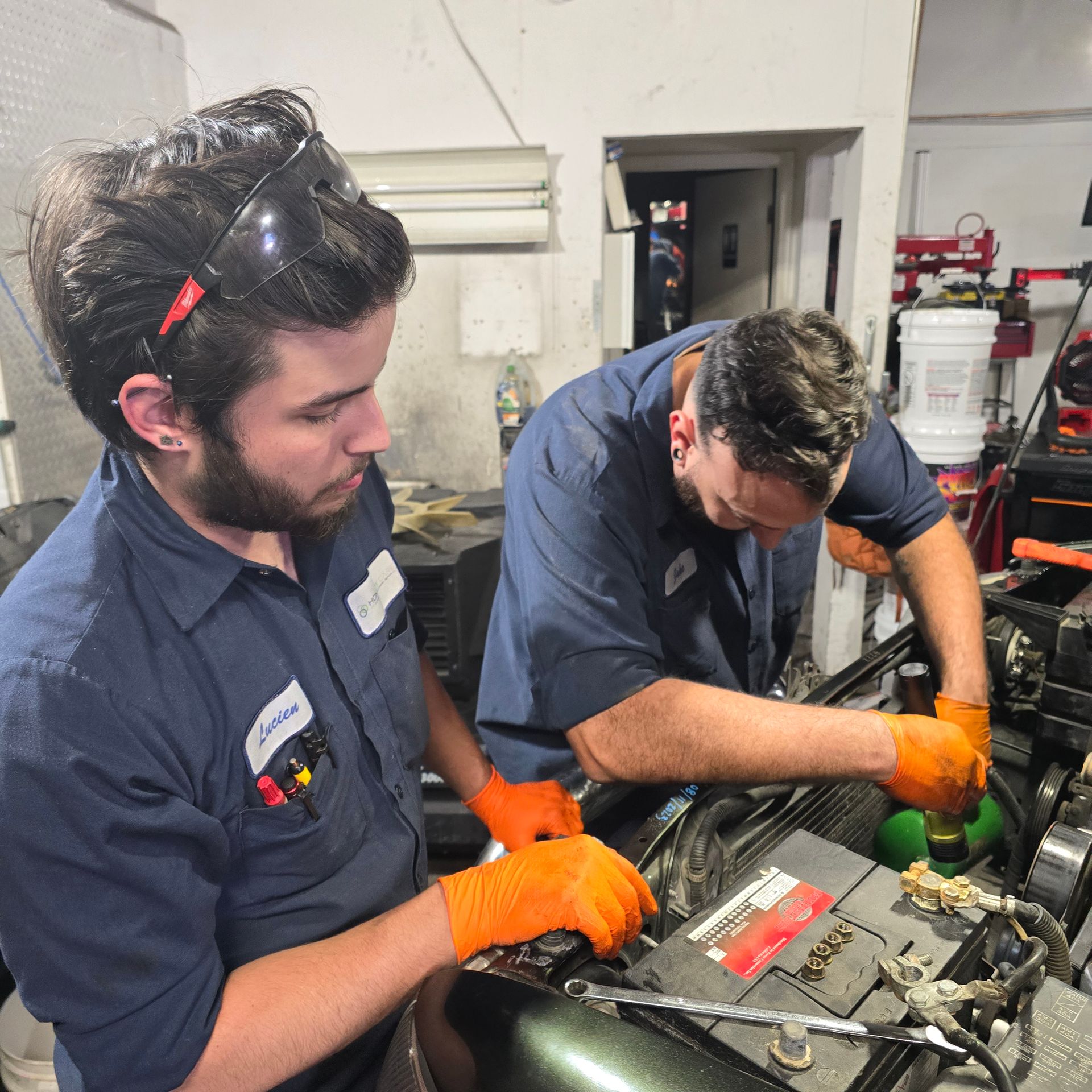 Two mechanics in blue shirts and orange gloves work on a car engine in a shop.