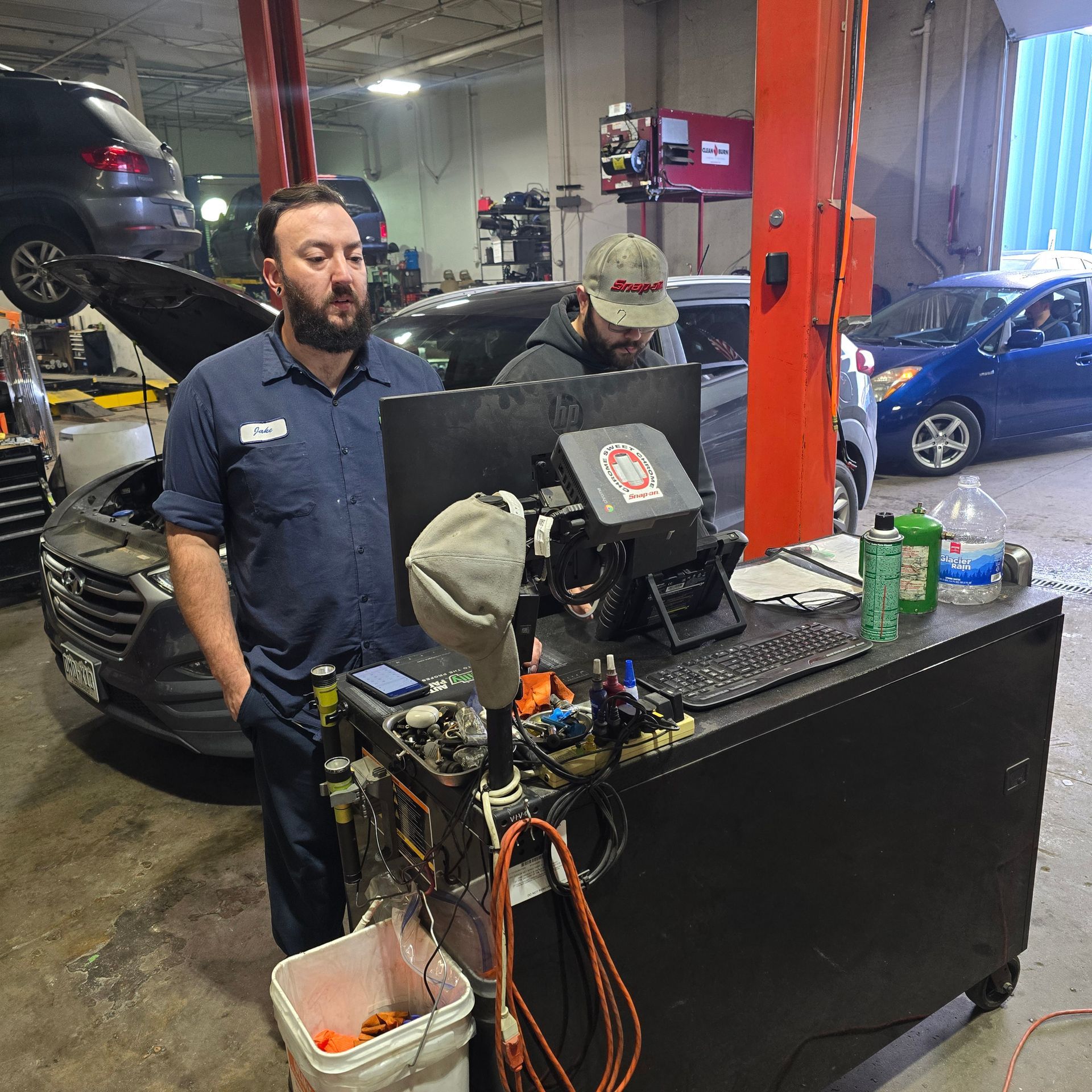 Two mechanics in a garage, one leaning, other at a computer. Cars being serviced, tools visible.