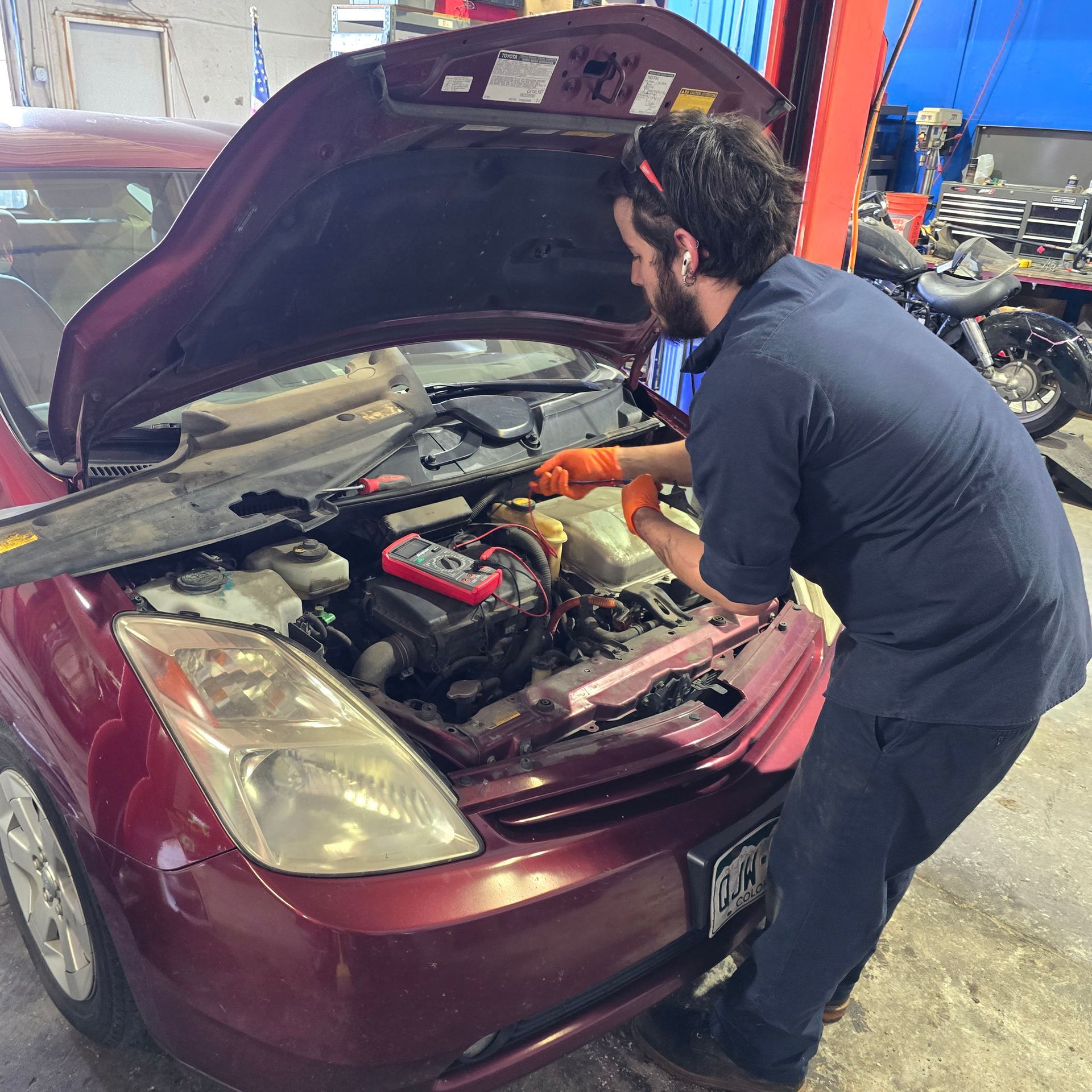 Mechanic working on a red car with the hood open, using a multimeter.