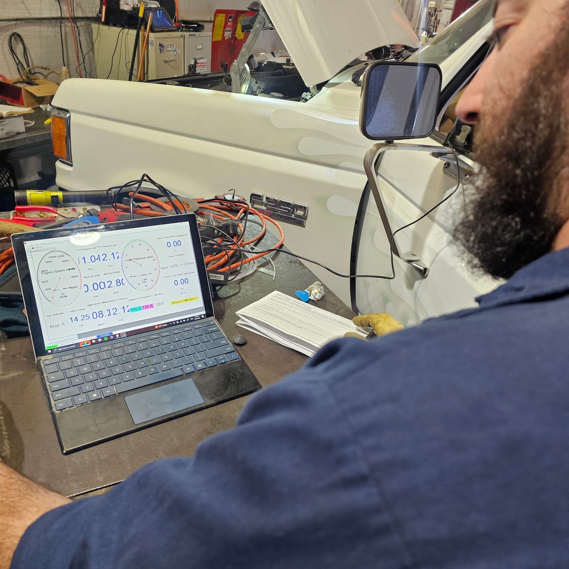 Mechanic using a laptop in a garage, vehicle in the background, wires and tools are scattered on the table.