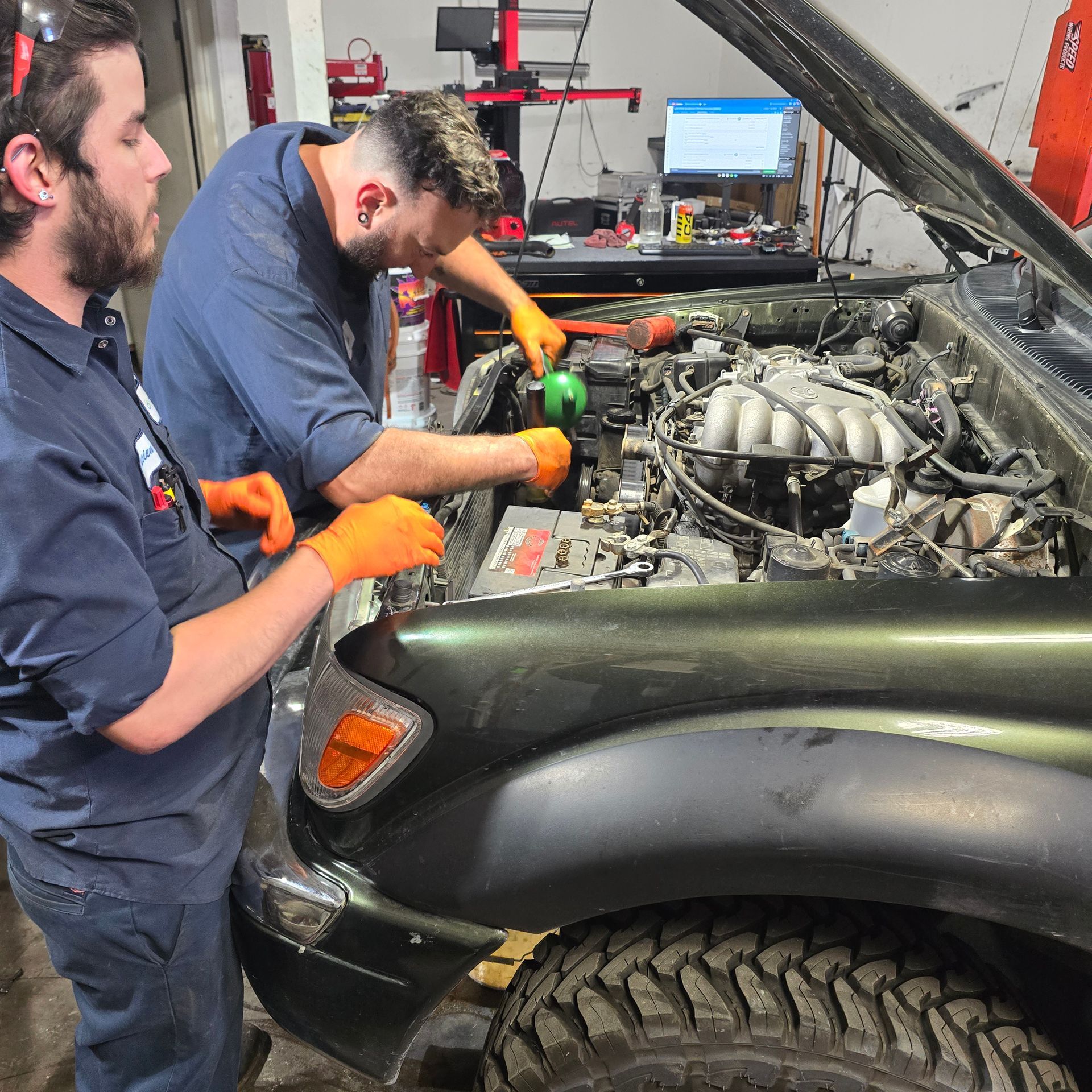 Two mechanics wearing gloves, working on an open car hood in a garage.