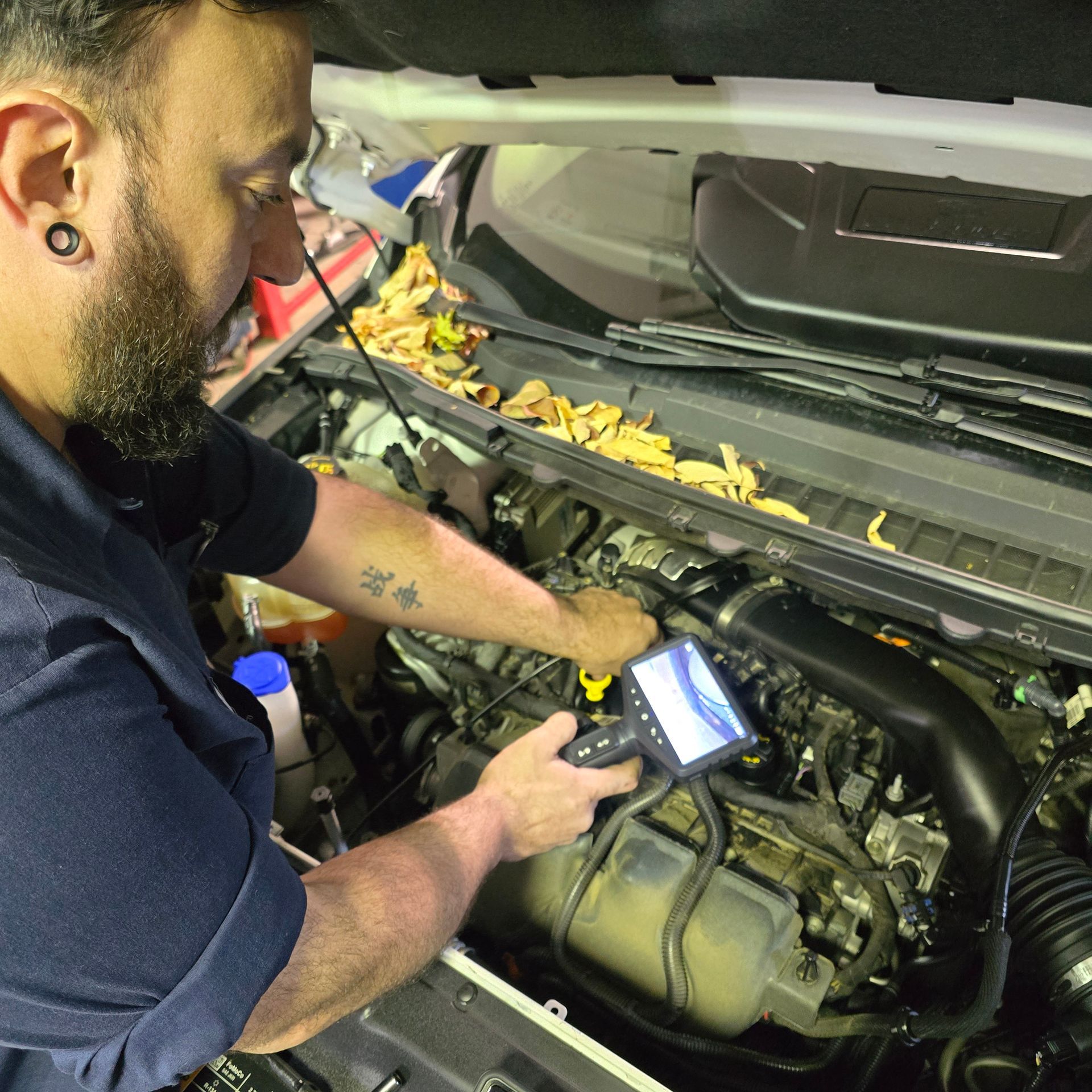Mechanic uses a diagnostic tool on a car engine in a garage; he’s wearing a black shirt and has tattoos.
