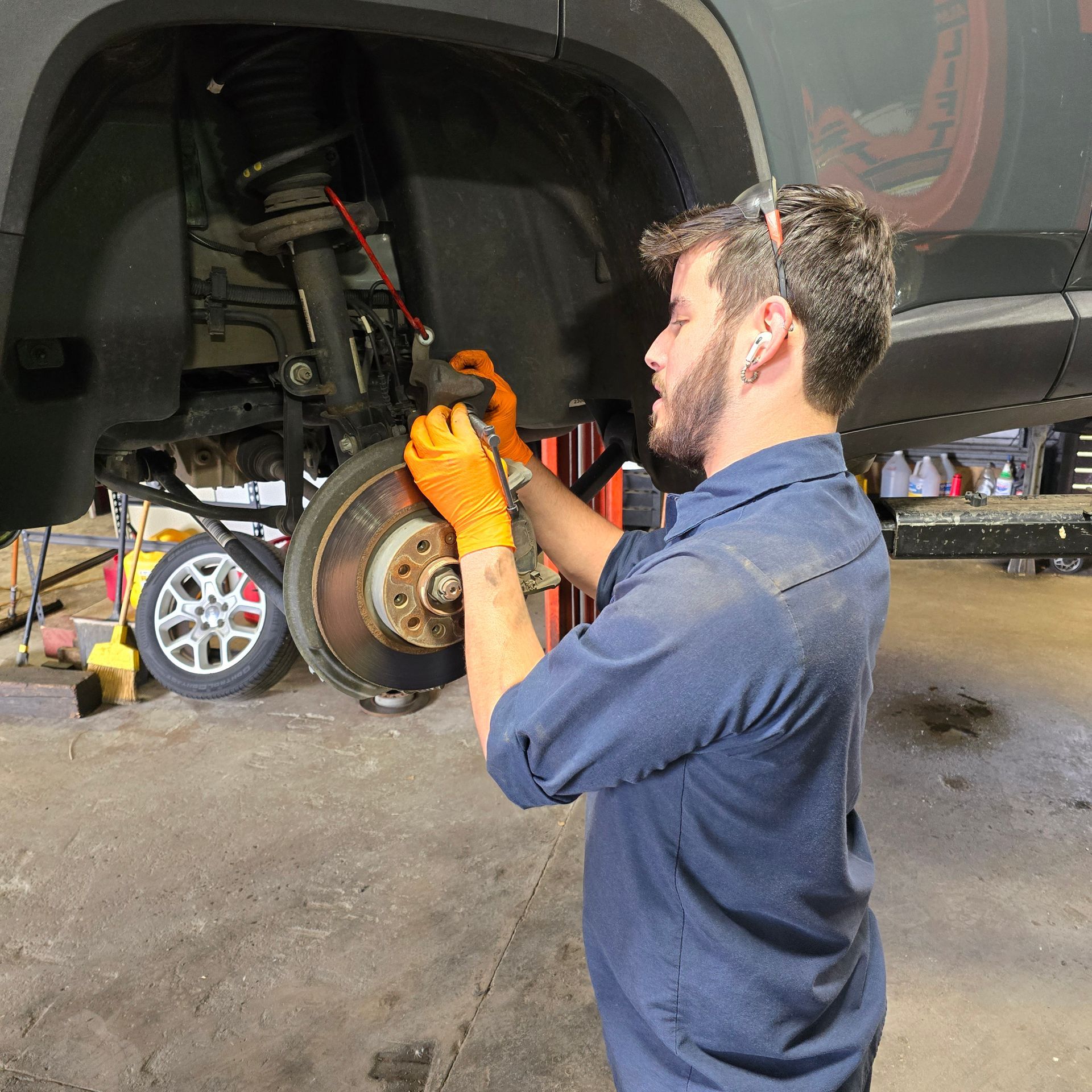 Mechanic working on car brakes; orange gloves, dark blue uniform, auto repair shop.