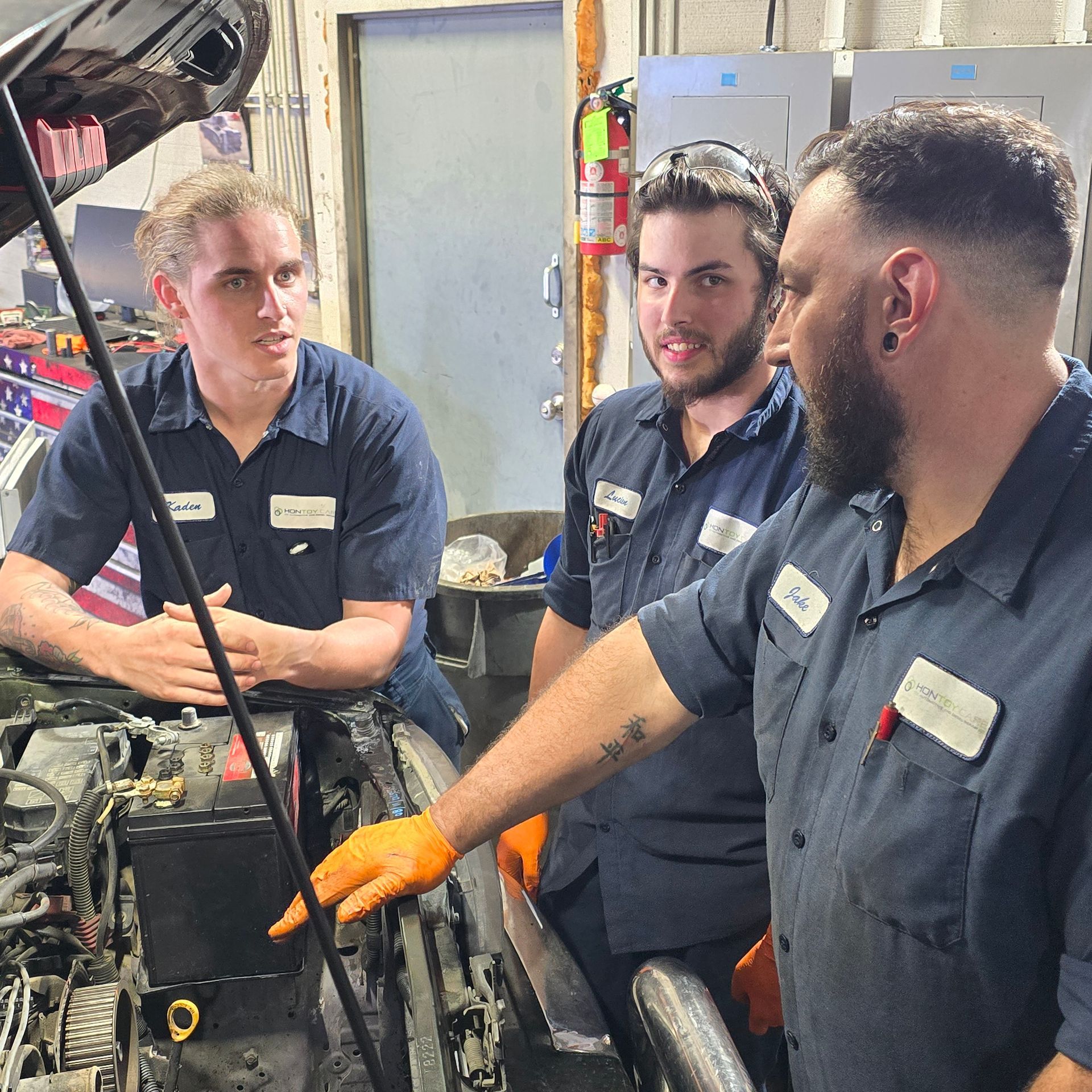Three mechanics in blue shirts examine a car engine in a garage; one points.