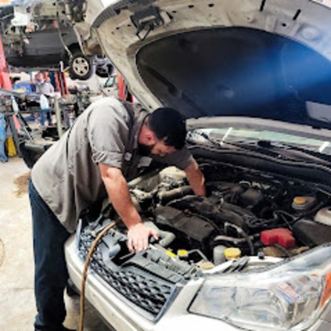 Mechanic working on a car engine in a garage; uses a tool, grey shirt, black cap.