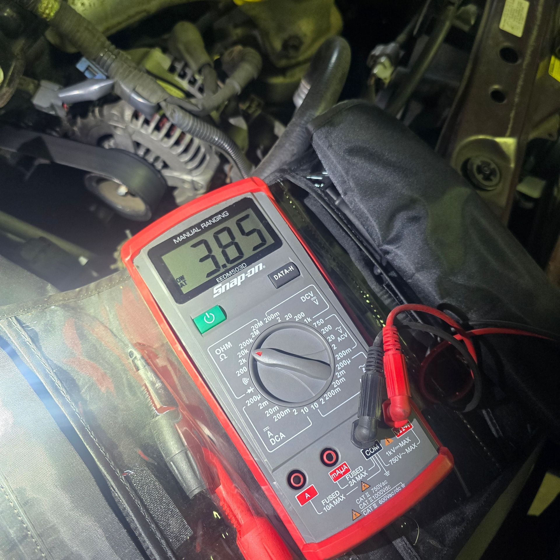 A Snap-on multimeter displaying 3.85 volts in an engine bay. Red and black test leads are attached.