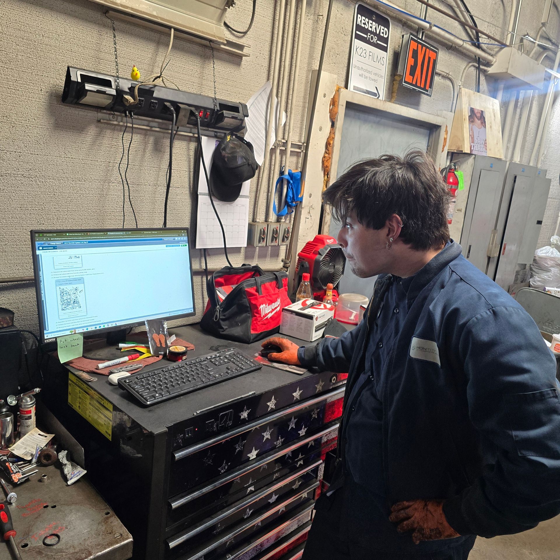 Mechanic at a computer in a garage. Wearing work clothes, looking at the screen, with a tool chest and equipment nearby.