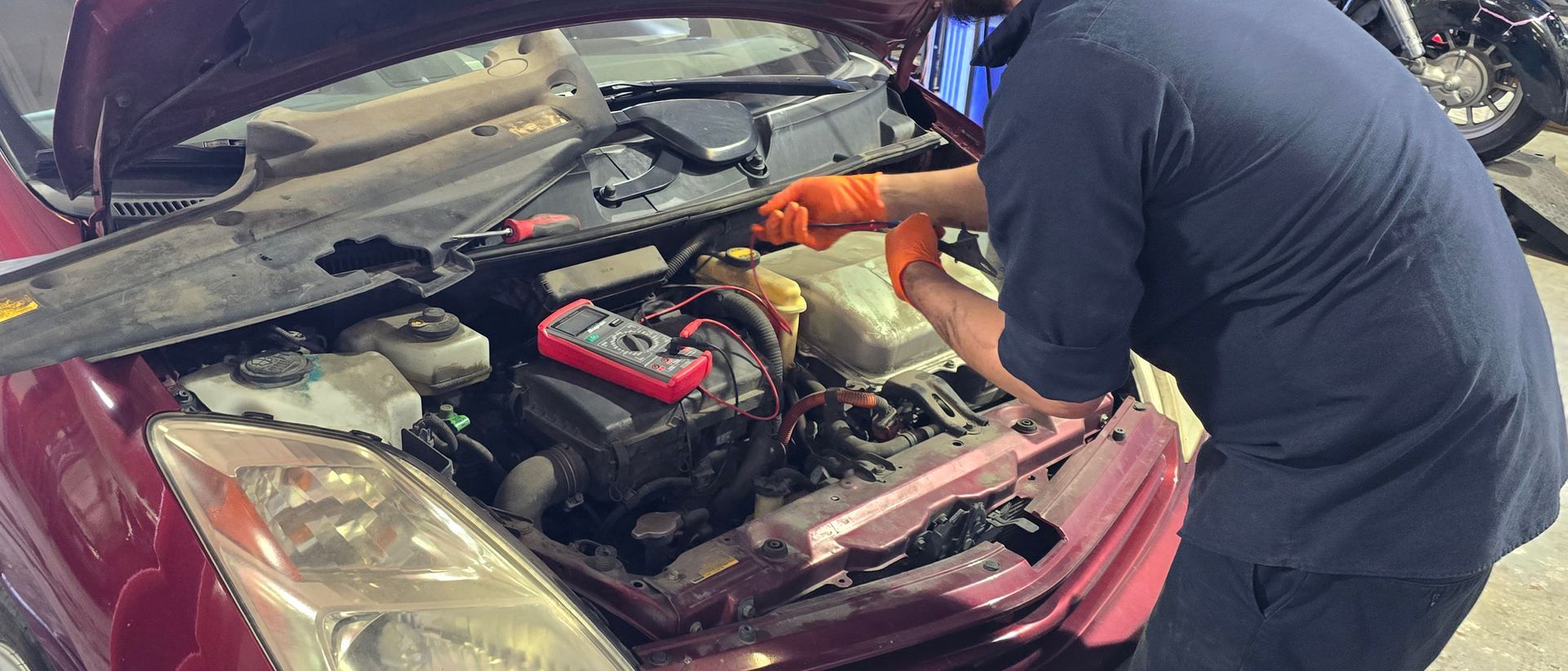 Person in blue shirt and orange gloves working on car engine, a red multimeter on top of the engine.