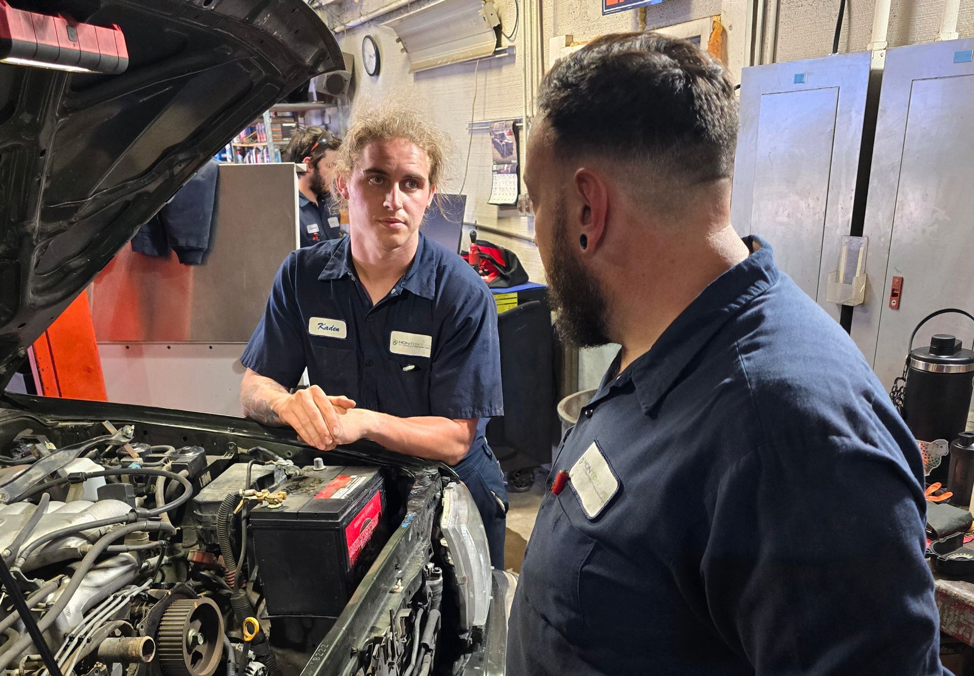 Two mechanics in blue shirts discuss an engine in a garage. One looks at the other.